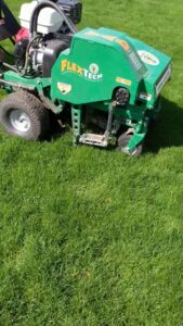 A green FlexTech lawn aeration machine actively working on a healthy lawn, provided by Nice & Precise Lawn Care in Ames, IA.