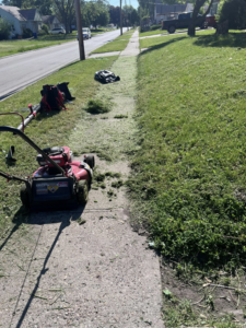 A lawnmower and leaf blower on a sidewalk with grass being mowed by Vick's Lawn Care Service in Des Moines, IA.