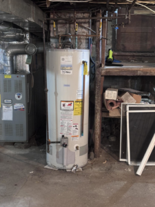 An old water heater showing signs of leakage and water damage in a basement, serviced by White Birch Plumbing in Derry, NH.