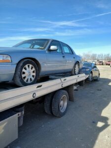 A light blue sedan loaded onto a flatbed tow truck, towing another car behind it, by JR's Services and Recovery, LLC in Indianapolis, IN.