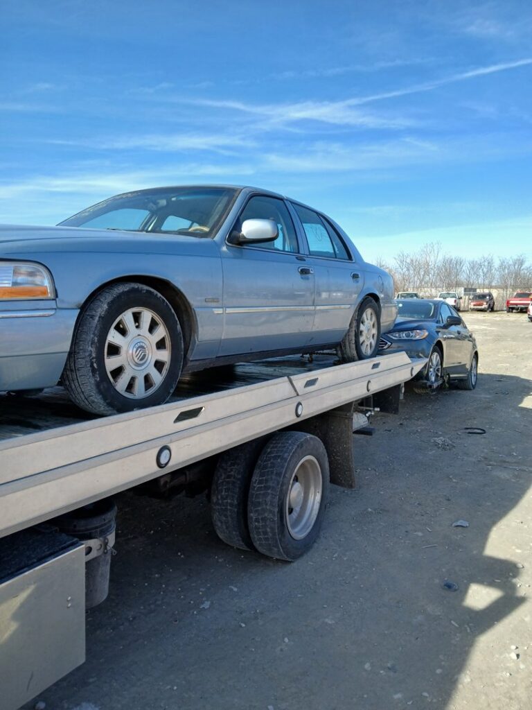 A light blue sedan loaded onto a flatbed tow truck, towing another car behind it, by JR's Services and Recovery, LLC in Indianapolis, IN.