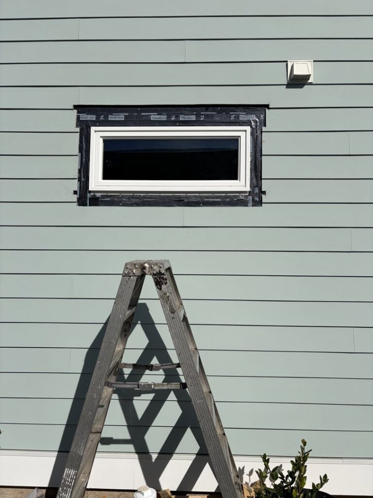 Close-up of light green horizontal siding installation around a window with a ladder, by Powell Roofing in Ladson, SC.