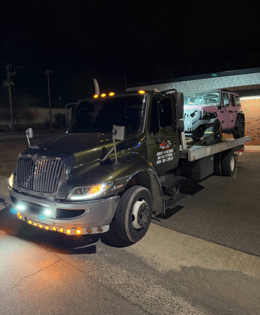 A light-colored Jeep loaded onto a flatbed tow truck from Bullcity Towing & Services LLC at night in Durham, NC.
