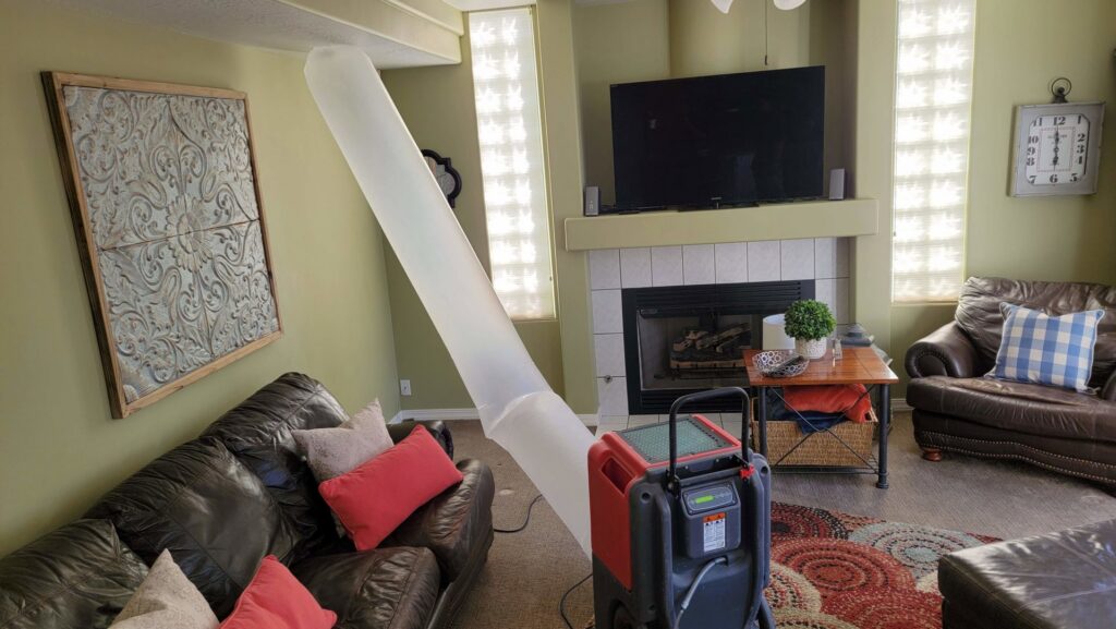 Professional drying equipment, including a large air duct and dehumidifier, set up in a living room for water damage restoration by Utah Disaster Team in Layton, UT.