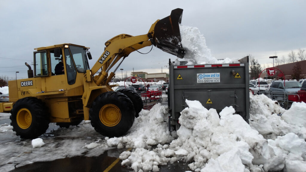 A yellow loader dumping snow into a large container in a parking lot for Snow & Ice Management Company in Pittsburgh, PA.