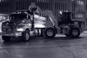 A front-end loader loading snow into a dump truck at night, demonstrating snow hauling services by JC Snow Management in Mount Vernon, NY