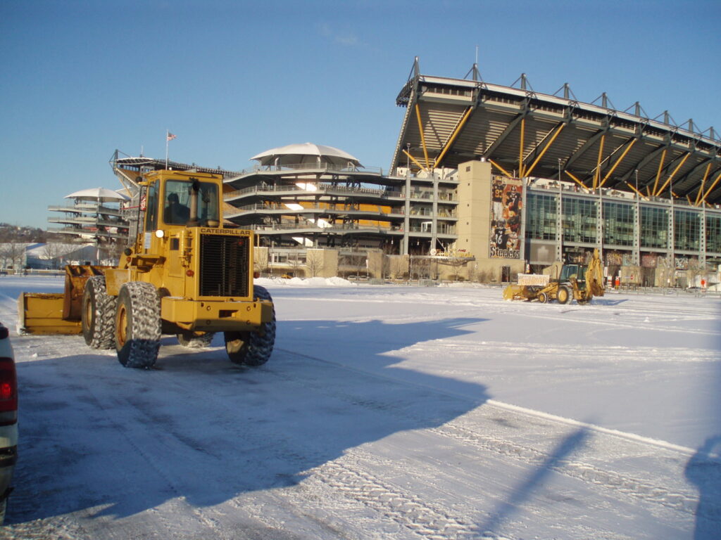 A large yellow loader plowing snow in a stadium parking lot for Snow & Ice Management Company in Pittsburgh, PA.