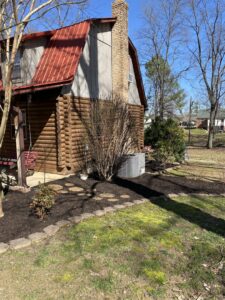 The side of a log cabin house with a well-maintained mulched garden bed around a bush and an AC unit by The Otherside Lawn Solutions in Ankeny, IA.