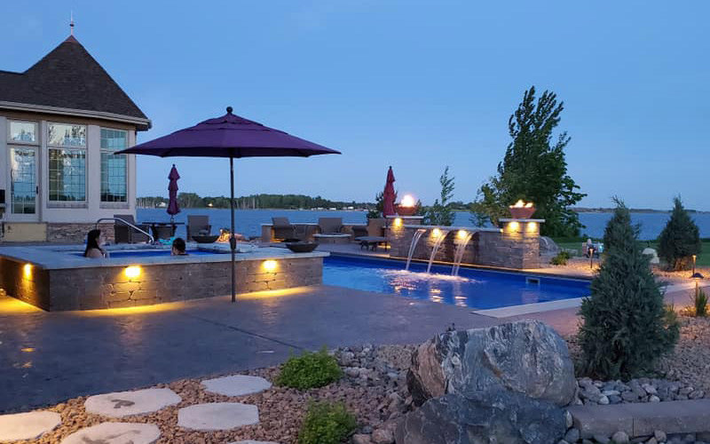 A luxurious illuminated pool and spa area with water features and fire bowls by Backyard Escapes by Kerner Pools & Spas in Bismarck, ND.