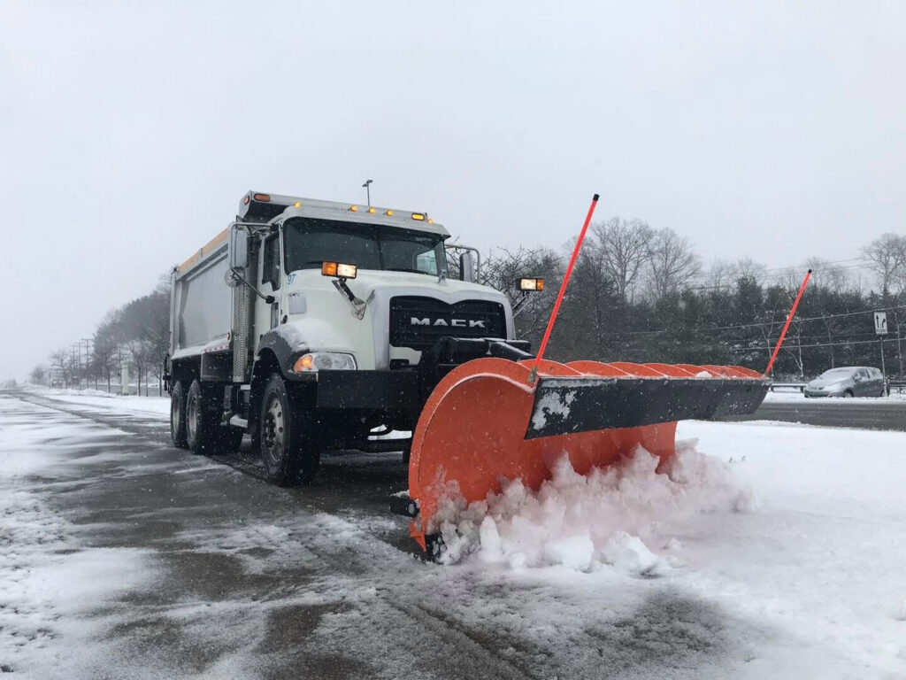 A large white Mack truck with an orange plow clearing snow from a road for Snow & Ice Management Company in Pittsburgh, PA.