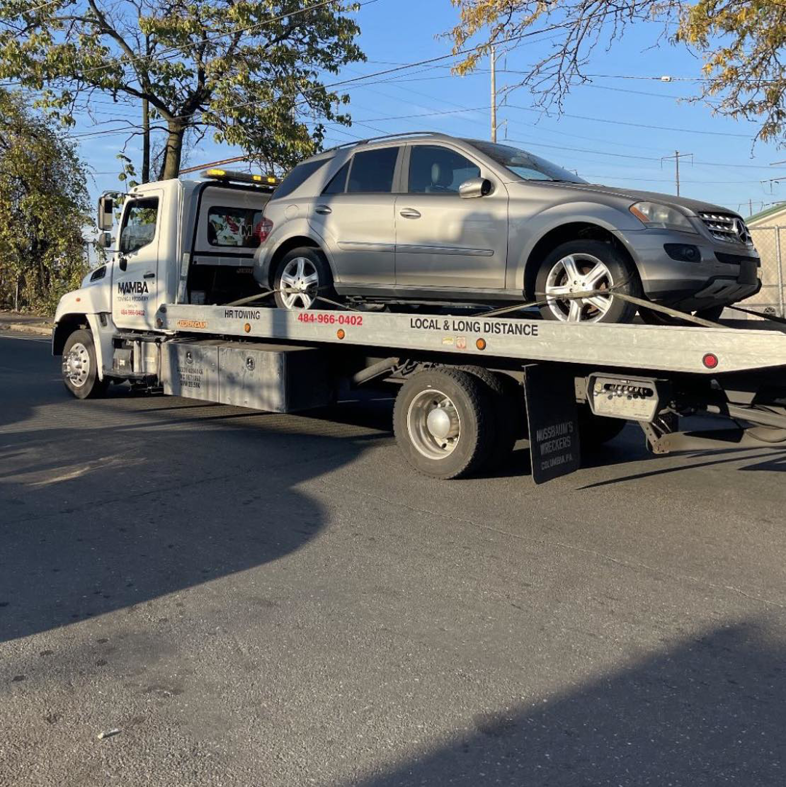 A Mamba Towing & Recovery flatbed truck towing a silver SUV on a street in Aurora, CO
