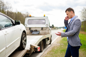 A man on the phone next to a white car being loaded onto a tow truck by DunRite Towing Jonesboro in Jonesboro, AR.