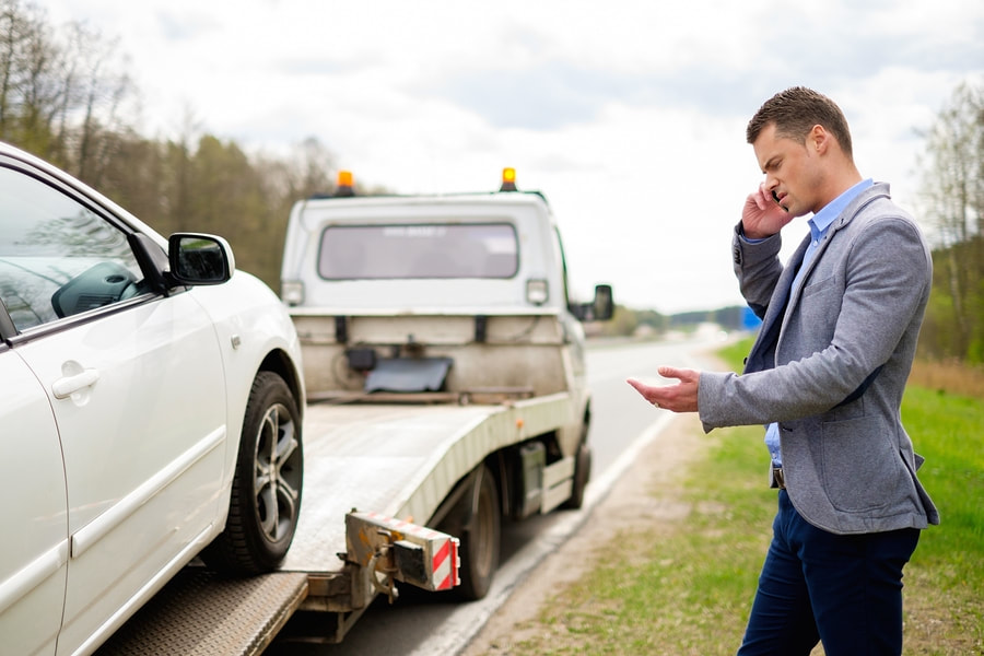 A man on the phone next to a white car being loaded onto a tow truck by DunRite Towing Jonesboro in Jonesboro, AR.