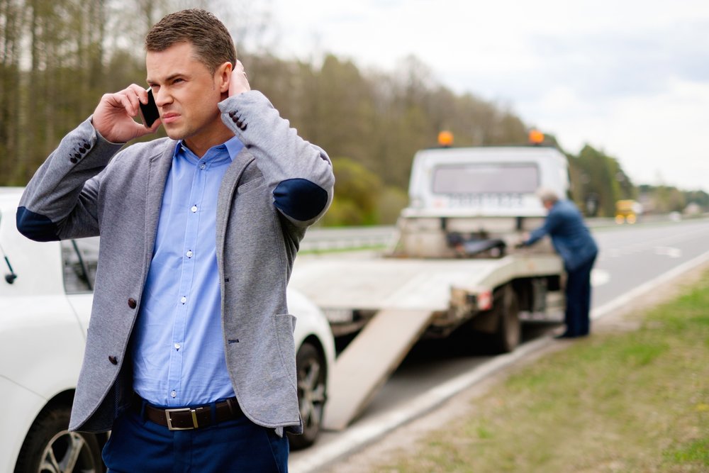 A man on the phone with a tow truck preparing to tow a white car on the roadside, representing Danny's Towing Edgar in Indianapolis, IN.