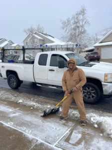 A man shoveling snow from a sidewalk next to a pickup truck, performing snow removal for Medina's Landscape Care LLC in Layton, UT.