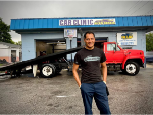 A man stands proudly in front of a red flatbed tow truck at Car Clinic Auto Repair in Fayetteville, NC, ready for towing services.
