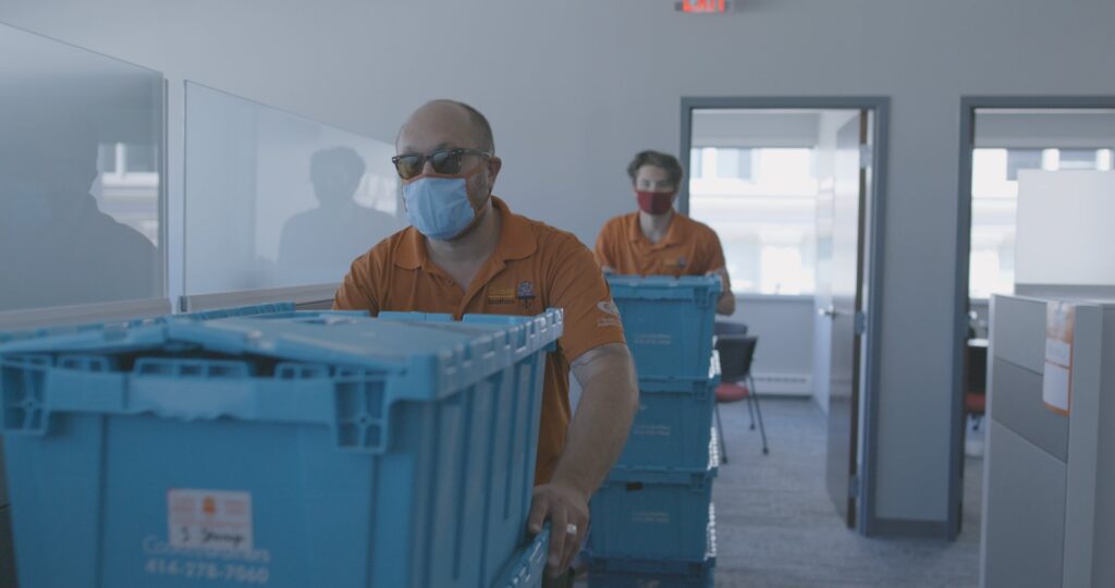 Two masked movers pushing blue moving crates in an office hallway for Coakley Brothers & Brothers Interiors in Milwaukee, WI.