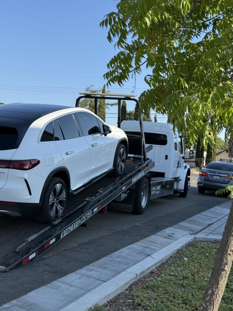 A white Mercedes-Benz EQE SUV being towed by an AJ's Towing Services & Roadside Assistance flatbed truck in Sacramento, CA.