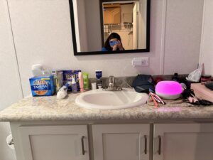 A messy bathroom vanity with various personal items, a 'before' scene for Heavenly Harlots Maid Cleaning Services in Grand Forks, ND.