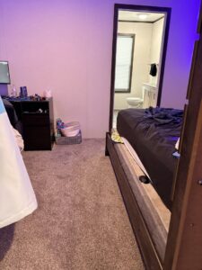 A messy bedroom with a visible bathroom doorway, showing a 'before' scene for Heavenly Harlots Maid Cleaning Services in Grand Forks, ND.