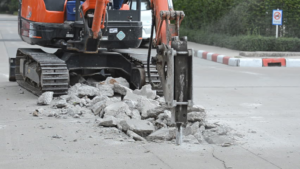 A mini-excavator with a hydraulic breaker attachment demolishing concrete on a road, a service by Torrado Demolition in Houston, TX.
