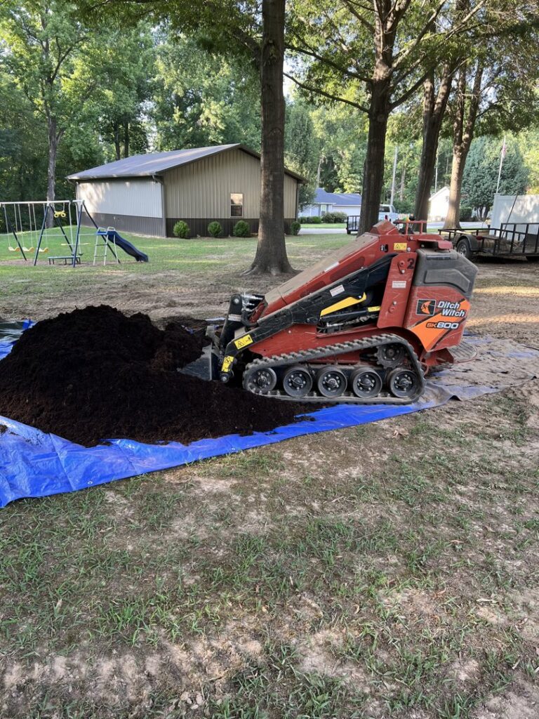 A mini skid steer loader efficiently moving a large pile of dark mulch for a landscaping project by The Otherside Lawn Solutions in Ankeny, IA.