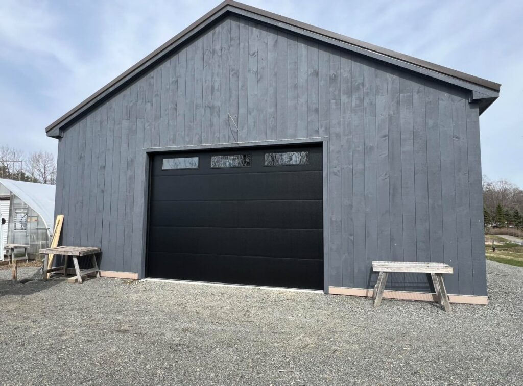 A modern black garage door installed on a gray wooden building by NDI Garage Door in Plaistow, NH.