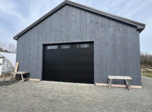 A modern black garage door installed on a gray wooden building by NDI Garage Door in Plaistow, NH.