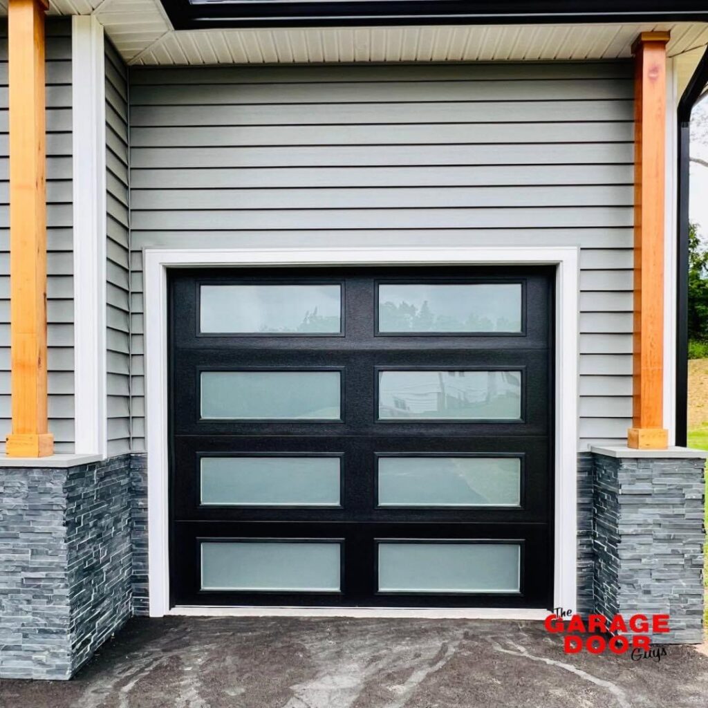 A modern black garage door with frosted glass panels installed on a grey-sided house by The Garage Door Guys, LLC in New Haven, CT.