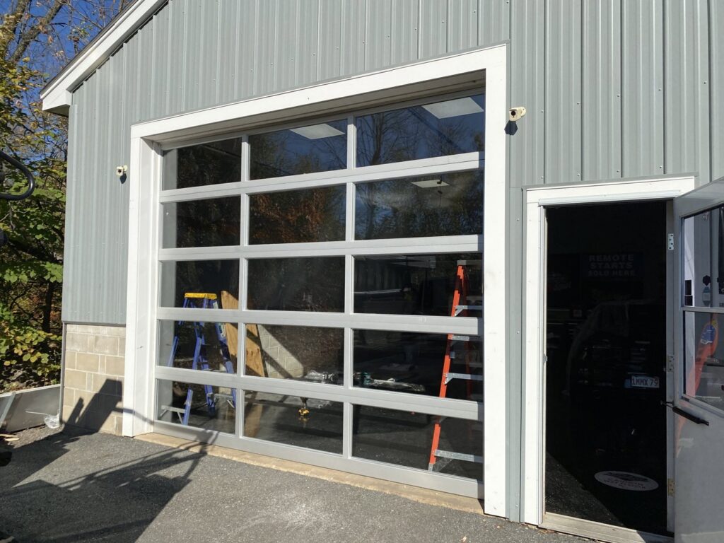 A modern full-view glass garage door installed on a building, showcasing work by Steve Shumsky Unlimited Overhead Doors LLC in Hudson, NH
