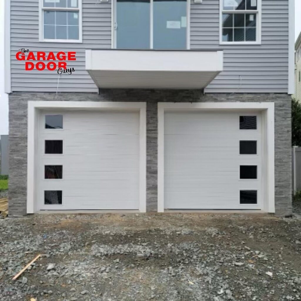 Two modern white garage doors with square windows installed on a grey-sided house by The Garage Door Guys, LLC in New Haven, CT.