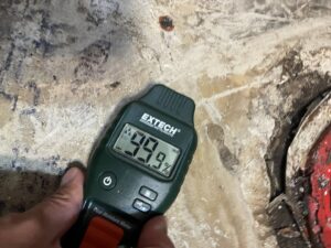 A moisture meter showing a 99.9% reading on a water-damaged floor, used by Roeun's Restoration and Cleaning LLC in Rochester, MN.
