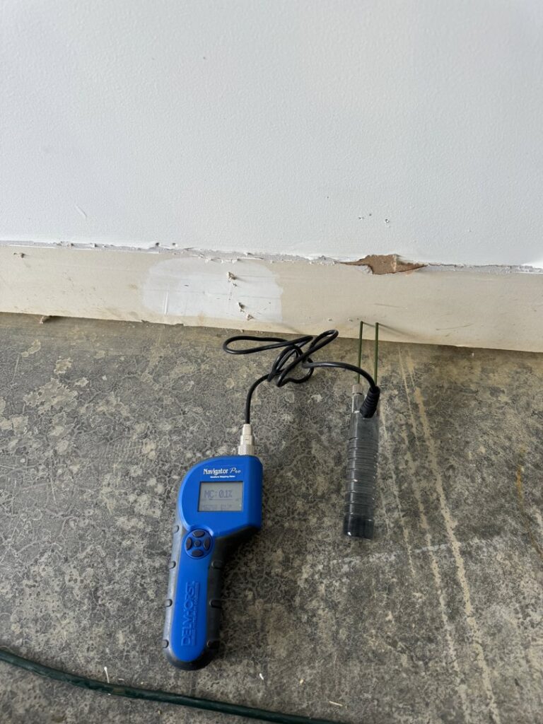 A moisture meter on a concrete floor next to a wall with visible water damage, used for assessment by KY Water Damage Restoration in Georgetown, KY.