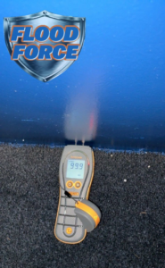 A moisture meter displaying '99.9 WET' on a carpeted floor, indicating water damage assessment by Flood Force in Dayton, OH.