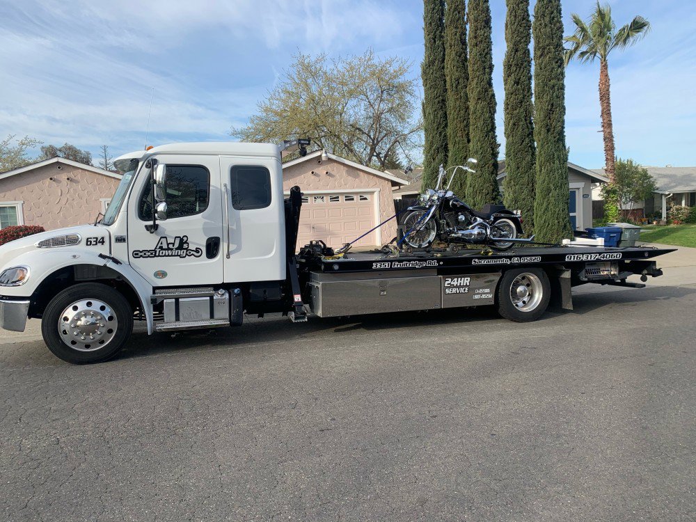 A white flatbed tow truck from AJ's Towing Services & Roadside Assistance towing a motorcycle in Sacramento, CA.