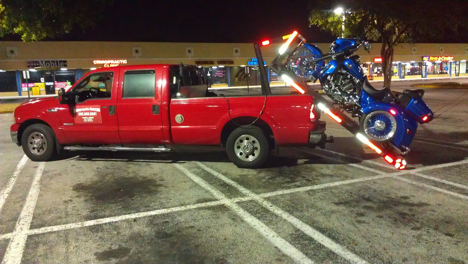 A red tow truck transporting a blue motorcycle at night, providing towing services by Motorcycle Rescue Inc. in Miami, FL.
