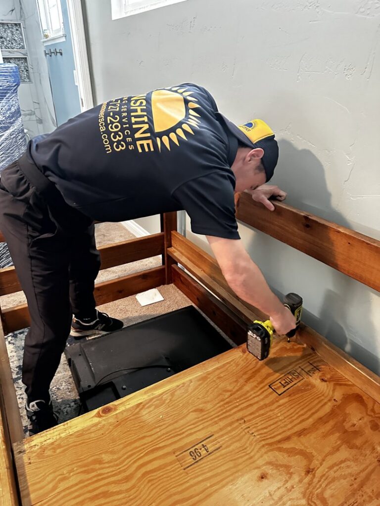 A mover from Sunshine Moving Services LLC assembling a bed frame with a drill, providing furniture services in San Diego, CA.