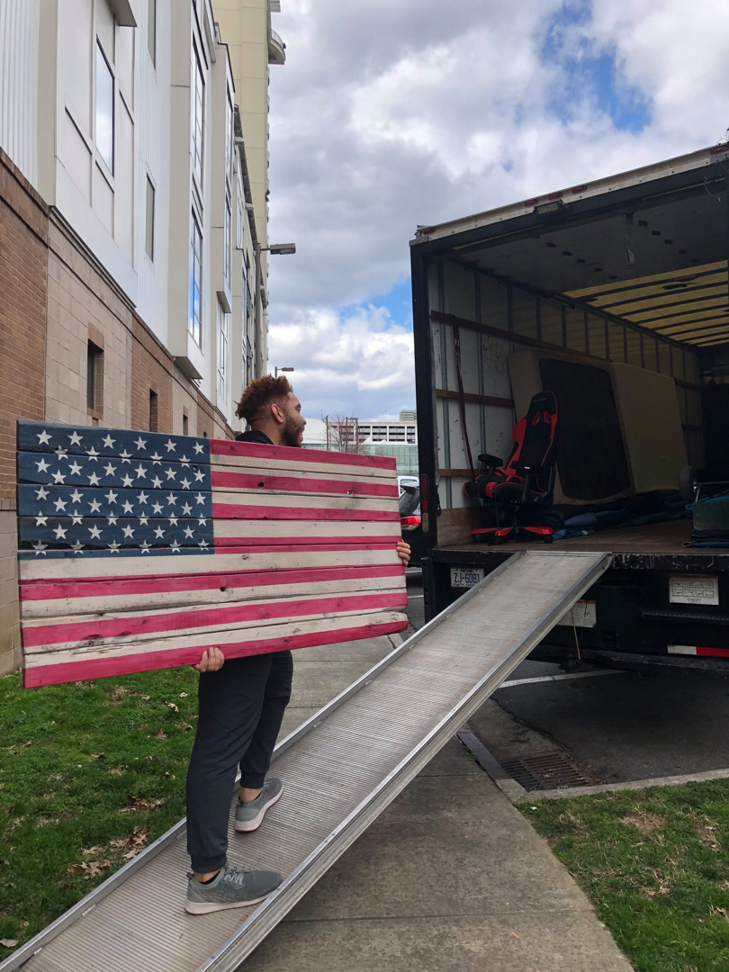 A Roma Moving team member carrying a large wooden American flag up a ramp into a moving truck in Charlotte, NC.