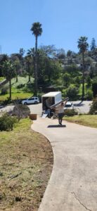 A Packman Moving professional carrying a long, wrapped item down a driveway towards a moving truck in San Diego, CA