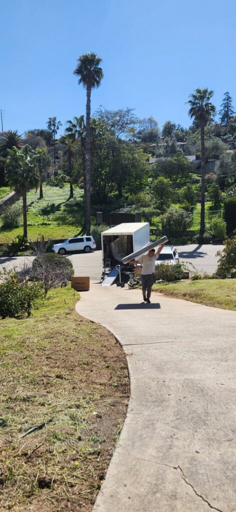 A Packman Moving professional carrying a long, wrapped item down a driveway towards a moving truck in San Diego, CA