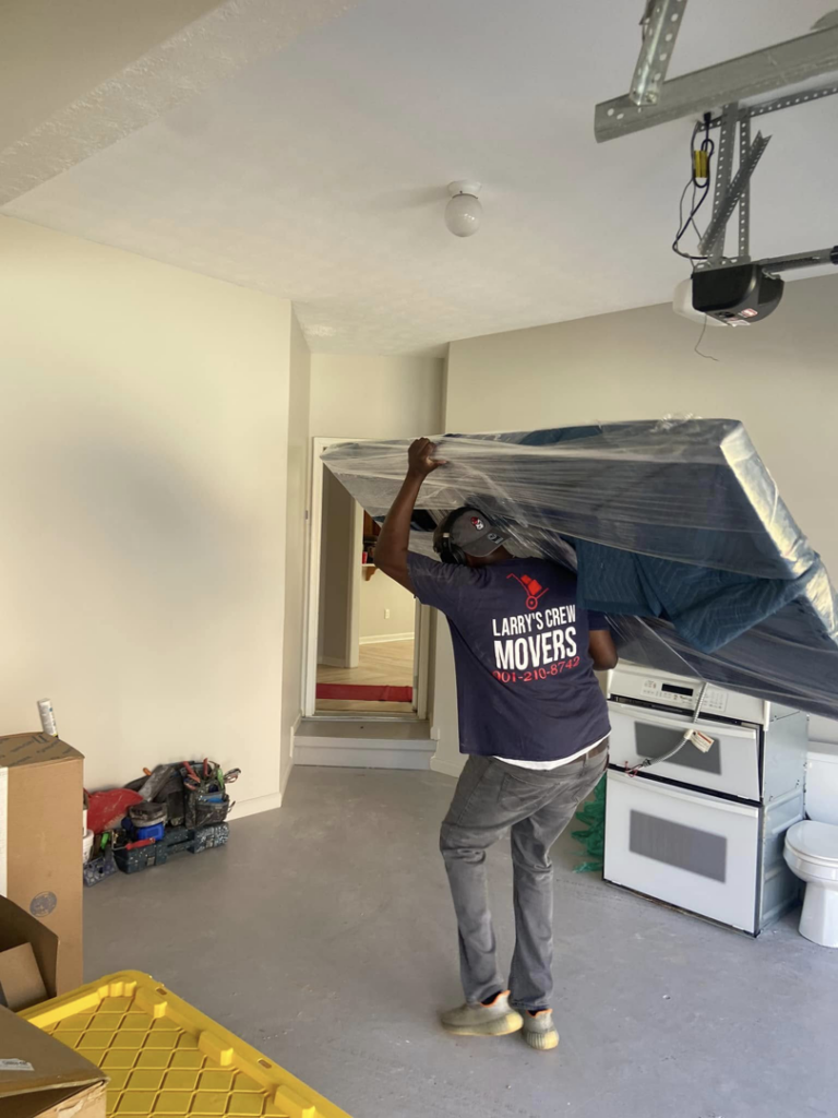 A mover from Larry's Crew Movers in Atlanta, GA, carrying a wrapped mattress into a garage.