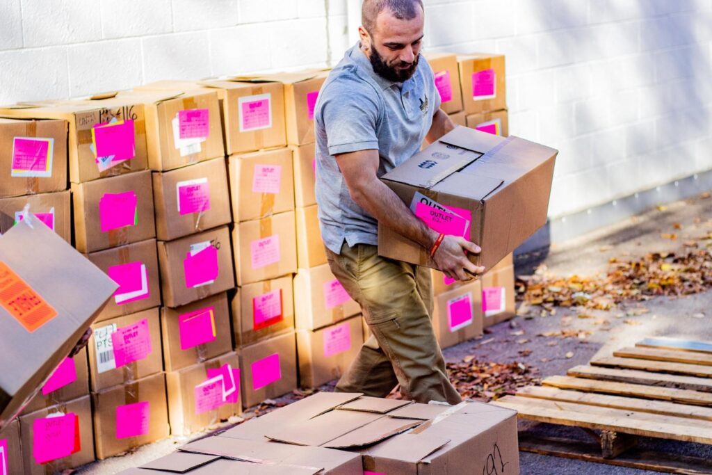 A PittMovers team member carrying a moving box outdoors, surrounded by other boxes in McKees Rocks, PA