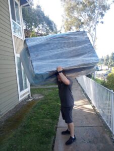 A Packman Moving professional carrying a large, shrink-wrapped piece of furniture in San Diego, CA