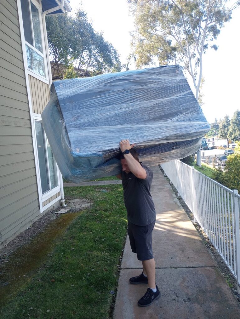 A Packman Moving professional carrying a large, shrink-wrapped piece of furniture in San Diego, CA