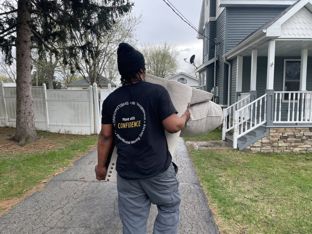A Shield Moving Services mover carrying a sofa cushion towards a house in Milwaukee, WI.