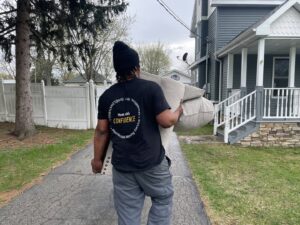 A Shield Moving Services mover carrying a sofa cushion towards a house in Milwaukee, WI.