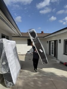 A mover carrying a large, wrapped mattress or board on his shoulder outside a home for All Way Moving in Washington, PA.