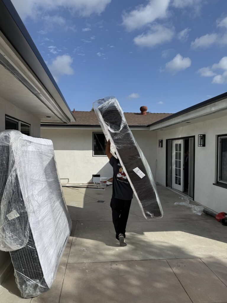 A mover carrying a large, wrapped mattress or board on his shoulder outside a home for All Way Moving in Washington, PA.