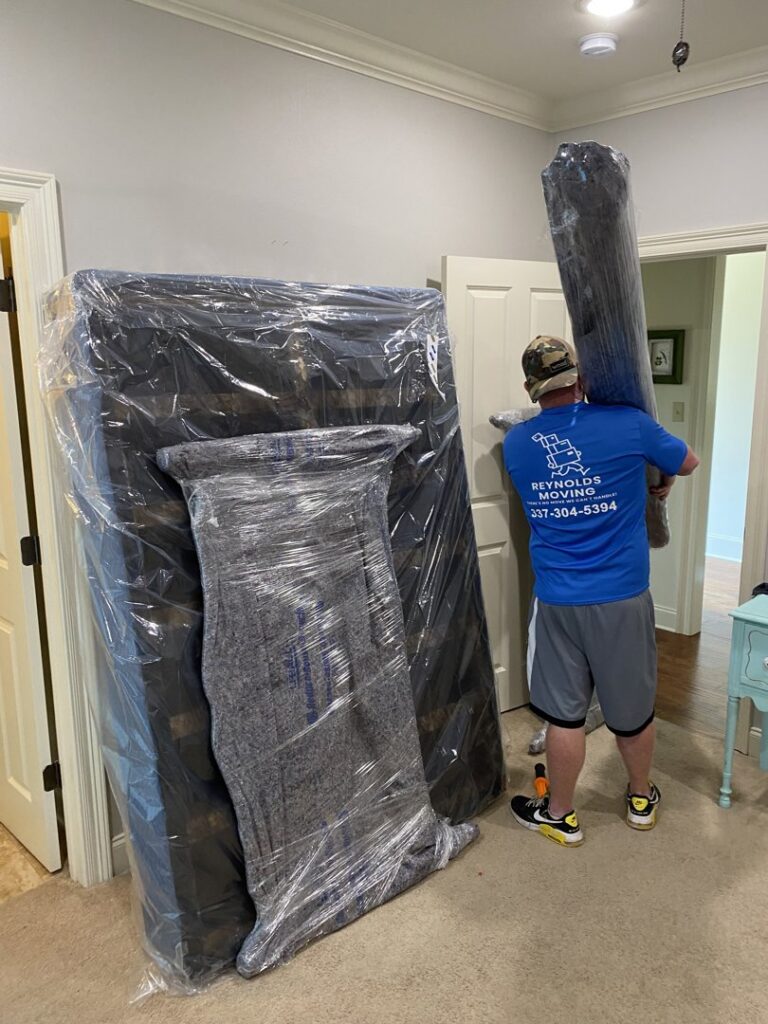 A mover from Reynolds Moving in Baton Rouge, LA, carrying a wrapped rug on his shoulder, with other packed items nearby