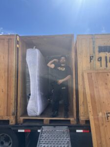 A Muscular Moving Men & Storage mover posing inside a large wooden crate on a truck with a wrapped mattress in Phoenix, AZ.